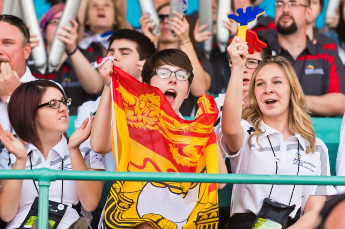 Event photo of students cheering for their home province of New Brunswick. Members from the crowd are waving a flag to show their team spirit, for the round of competitor introductions during the opening ceremony.