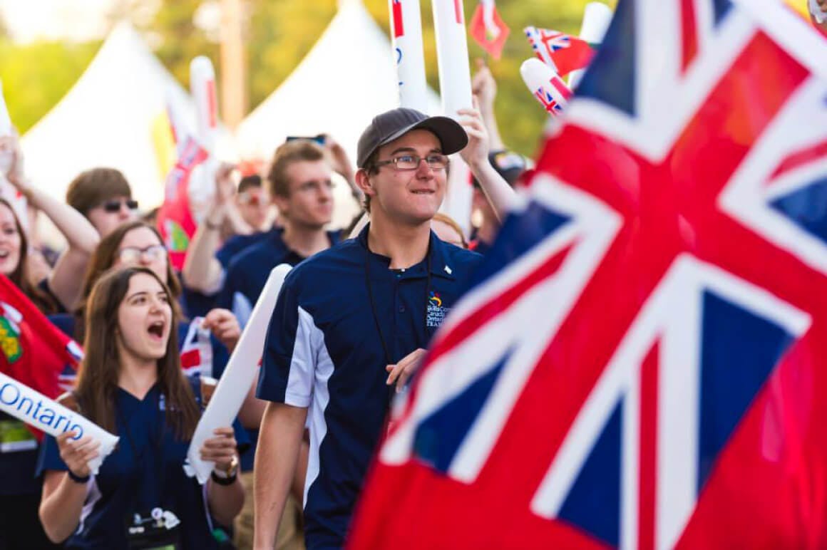 Event photo of students and teachers from the province of Ontario showing their team spirit by holding up their provincial flags. They are seen marching towards the main stage during the opening ceremony.